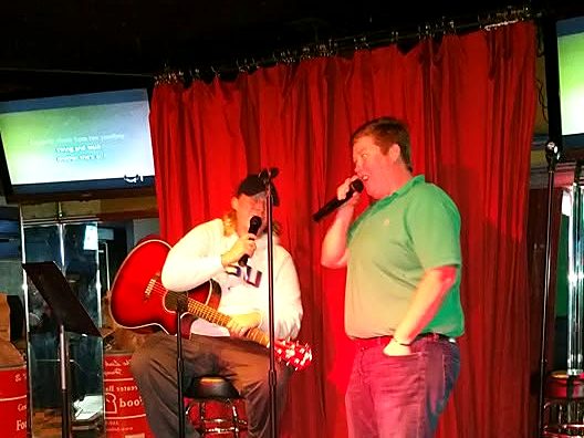 Hollie playing guitar and singing on a bar stage with another singer
